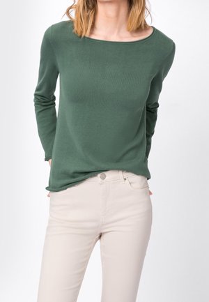 Vrouw met een groen shirt met lange mouwen, licht ingestopt in een hooggetailleerde, crèmekleurige broek, met de handen achter haar rug, voor een witte achtergrond.