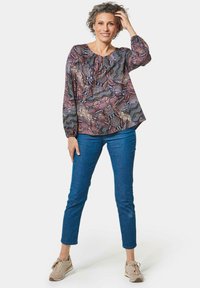 Langärmlige gemusterte Bluse in Lila- und Rosatönen, mit lockerer Passform und gerafftem Ausschnitt. Kombiniert mit enganliegenden blauen Jeans und beigen Schuhen.