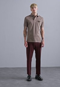 Polo marron avec un col, logo brodé sur la poitrine, associé à un pantalon bordeaux foncé et des chaussures noires, se tenant sur un fond gris.