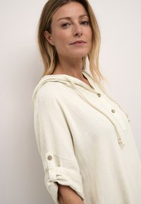 Pullover con cappuccio di un beige chiaro realizzato in un tessuto morbido, con una chiusura frontale con bottoni, polsini arrotolati e dettagli con lacci. Texture liscia.