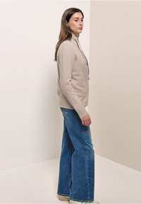 Nainen seisoo sivuttain beige vetoketjullisessa neuletakissa, leveälahkeisissa sinisissä farkuissa ja beigeissä kengissä neutraalin taustan edessä.