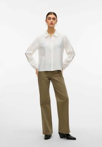 Camicia bianca a maniche lunghe con bottoni, realizzata in un tessuto texture, abbinata a pantaloni a gamba larga di colore verde oliva. Stivali neri con tacco completano l'outfit.