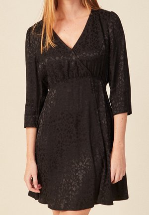 Vrouw draagt een zwarte jacquard wikkeljurk met bloemenprint, driekwart mouwen en kleine stoffen knopen op de taille.