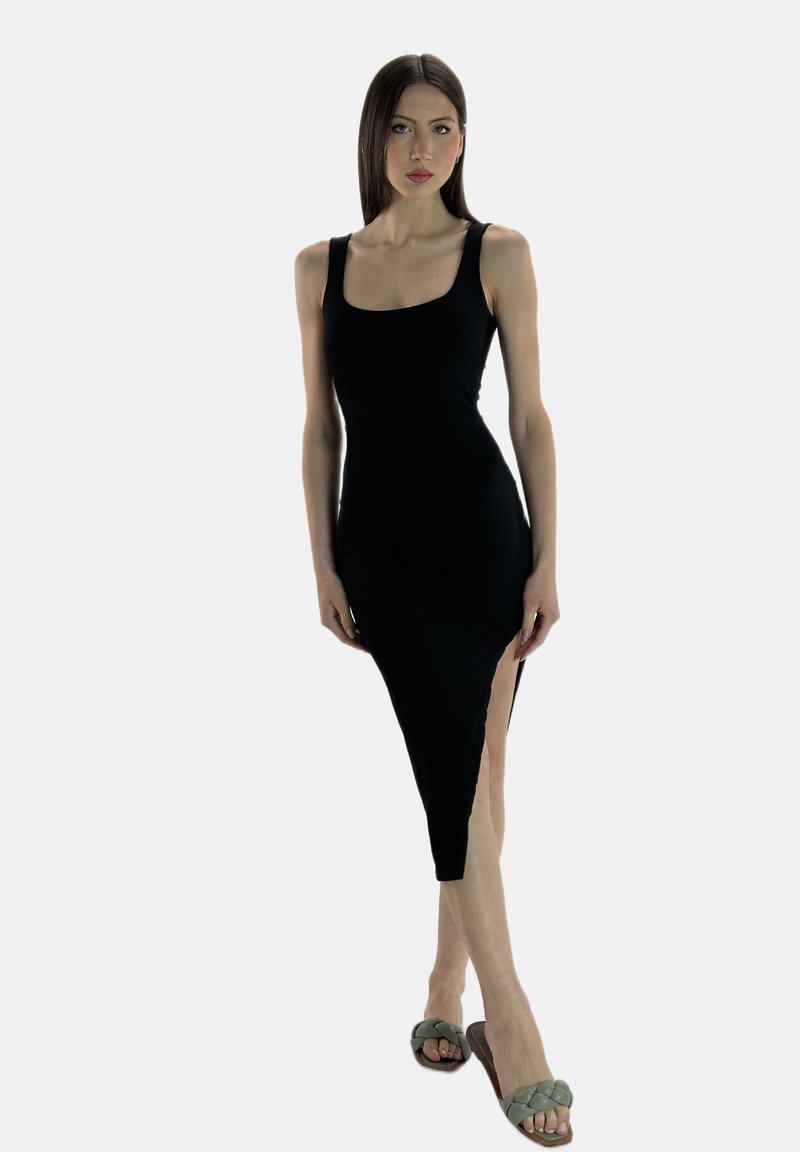 Schwarzes ärmelloses Kleid mit taillierter Passform und seitlichem Schlitz; getragen mit grünen geflochtenen Sandalen. Glatte Textur, figurbetonte Silhouette.