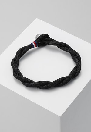 Zwart gevlochten touwarmband met zilveren sluiting en een klein detail van rood, wit en blauw draad, weergegeven op een wit schuin oppervlak.