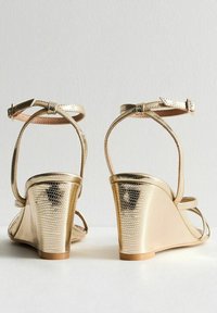 Sandalias de cuña doradas con un acabado texturizado similar a la piel de un reptil. Cuentan con correas en el tobillo, un hebilla segura y una suela suave y curva.