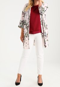 Kimono léger à imprimé floral en rose pâle, doté de manches trois-quarts, d'un devant ouvert et d'un bord noir, porté sur un haut rouge et un pantalon blanc.