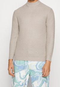 Beige stickad tröja med lång ärm, kombinerad med ljusa byxor med abstrakt mönster i blå, grön och vit färg.