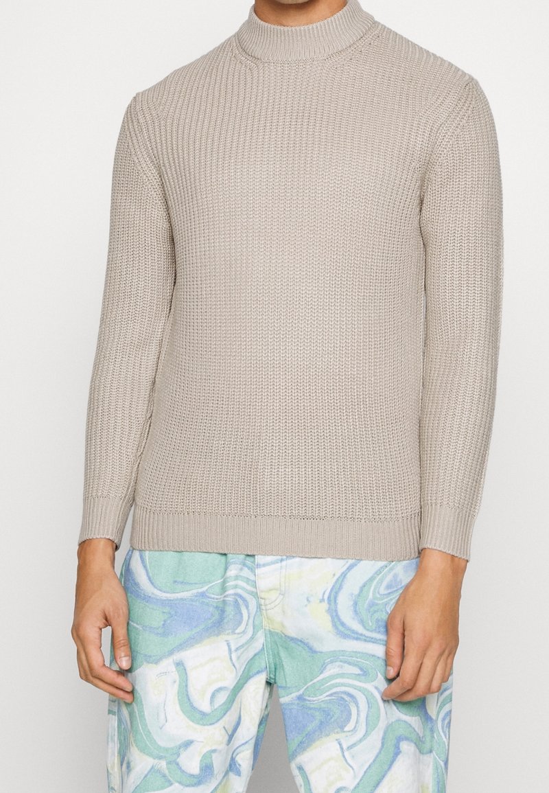 Beige stickad tröja med lång ärm, kombinerad med ljusa byxor med abstrakt mönster i blå, grön och vit färg.