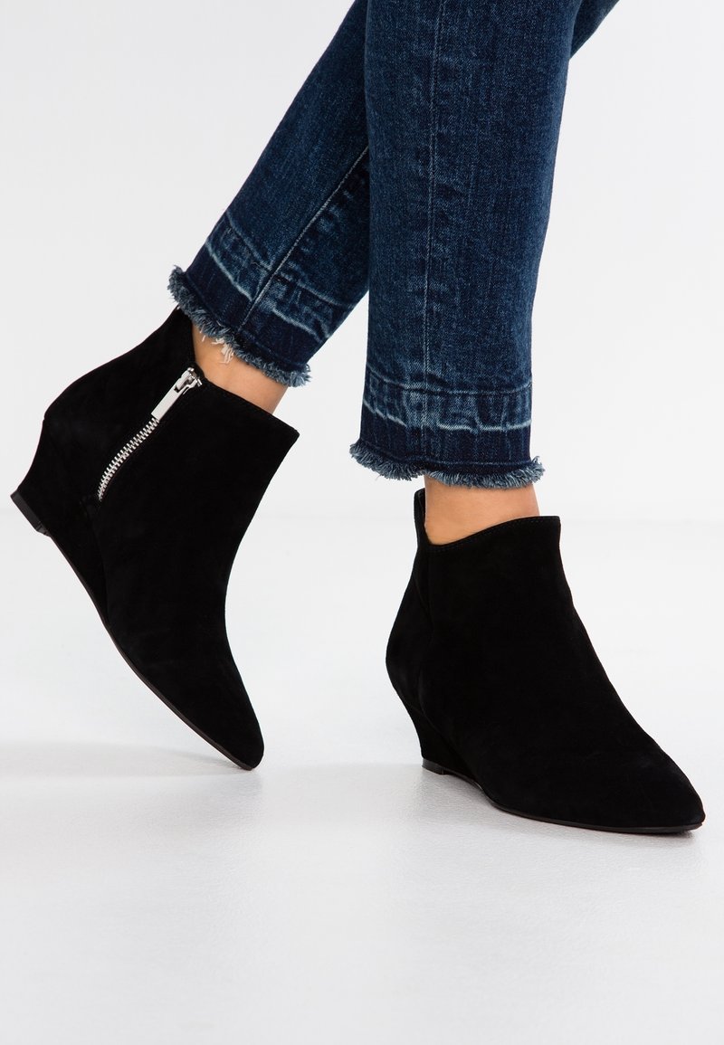 Bottines à talon en daim noir avec un bout pointu et une fermeture éclair sur un côté, associées à un jean en denim foncé avec un ourlet effiloché.