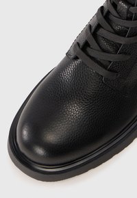 Bota de cuero negro con un acabado texturizado, punta redonda y suela de goma negra resistente. Presenta cordones negros a juego y detalles de diseño minimalistas.