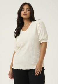 Weißes gestricktes Oberteil mit V-Ausschnitt, kurzen Puffärmeln, strukturiertem Muster und gewelltem Saum. Kombiniert mit schwarzen Hosen.
