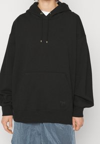 Sudadera con capucha negra confeccionada con tela suave, que incluye un bolsillo delantero espacioso, cordones ajustables con extremos de metal dorado y un parche con el logo.