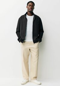 Muž v čiernej bunde na zips cez bielu košeľu, béžových nohaviciach a bielych teniskách, stojaci s rukami v kapse na bielom pozadí.