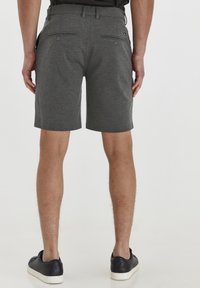 Man wearing dark gray tailored shorts with buttoned back pockets and black sneakers, standing on a white background.