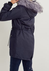 Parka bleu marine avec capuche en fausse fourrure, taille resserrée par cordon, poches latérales et manches élastiques. Matière lisse avec une finition légèrement brillante.