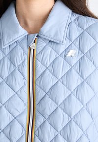 Veste matelassée bleu clair avec un col plié, une fermeture éclair rayée et un logo blanc sur la poitrine, portée par une personne aux cheveux longs.
