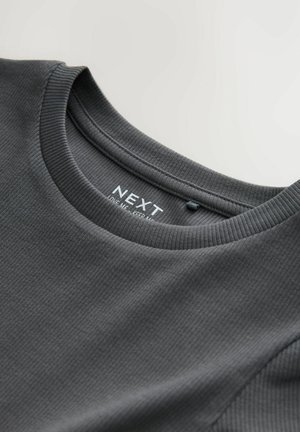 T-shirt a maniche corte color grigio scuro, con una texture morbida; presenta un'etichetta con il logo "NEXT". Il colletto ha una finitura liscia.