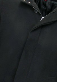 Zwarte jas met gedeeltelijk open rits over een met knopen gesloten klep, waarbij de binnenvoering zichtbaar is.