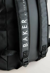 Sac à dos noir avec des bretelles rembourrées, présentant une finition texturée et un logo "BAKER" blanc. Comprend un matériel de boucle ajustable et des poches latérales.