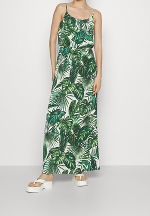 Mujer con vestido largo sin mangas con estampado de hojas tropicales verdes y sandalias blancas de plataforma, posando frente a un fondo liso.