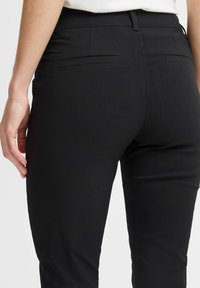 Schwarze Hose mit glatter Textur, ausgestattet mit zwei Gesäßtaschen und einer figurbetonten Passform, seitlich und von hinten gezeigt.