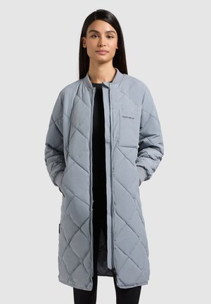 Frau trägt einen langen, hellgrauen Steppmantel mit Reißverschluss vorne und den Händen in den Taschen vor einem schlichten Hintergrund.