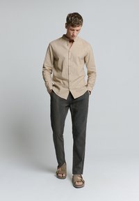 Beige langarm shirt zonder kraag, gecombineerd met donkergrijze broeken en brown sandalen, die een ontspannen pasvorm en casual stijl laten zien.