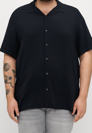 Mężczyzna w luźnej, czarnej koszuli z krótkim rękawem i guzikiem oraz szarych spodniach, z widocznymi tatuażami na lewym przedramieniu, stoi na jednolitym tle.