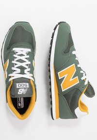 Sneaker verde e grigio con accenti gialli, tessuto in rete, lacci bianchi e suola in gomma nera. Etichetta "500" sulla linguetta.