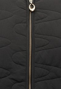 Μαύρο καπιτονέ ύφασμα με ένα κεντρικό χρυσό φερμουάρ και στρογγυλεμένα, γλυπτά σχέδια ραφής σε κάθε πλευρά.