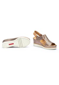 Sandalias de cuña de bronce con diseño superior perforado, punta abierta y tirante en el talón. Cuenta con una suela exterior de goma blanca texturizada con patrón en zigzag.