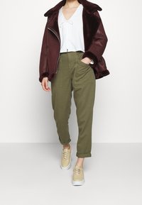 Giacca in shearling bordeaux sopra una camicetta bianca, abbinata a pantaloni verde oliva e sneakers beige con suola bianca.