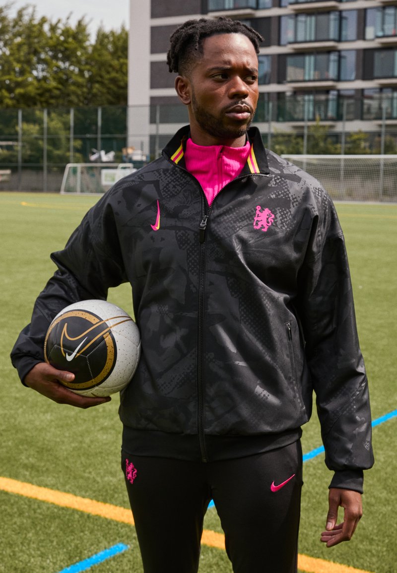Giacca nera con zip, dettagli rosa e motivo camouflage, abbinata a pantaloni neri con logo rosa. Tiene un pallone da calcio bianco e nero.