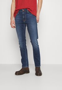 Kape slim-fit tummansiniset farkut, joissa on hienovarainen haalistuminen, oranssit ompeleet ja viisi taskua, yhdistettynä ruskeisiin nahkasaappaisiin.