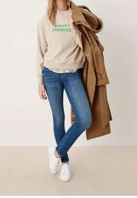 Sweat-shirt beige avec le texte « positive MINDSET », superposé à une chemise à fleurs, associé à un jean bleu et des baskets blanches. Manteau marron porté sur le bras.