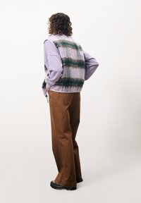 Übergroßer lila Pullover, der unter einer grün-beigen karierte Weste getragen wird, kombiniert mit braunen weit geschnittenen Hosen und schwarzem Schuhwerk. Rückansicht.