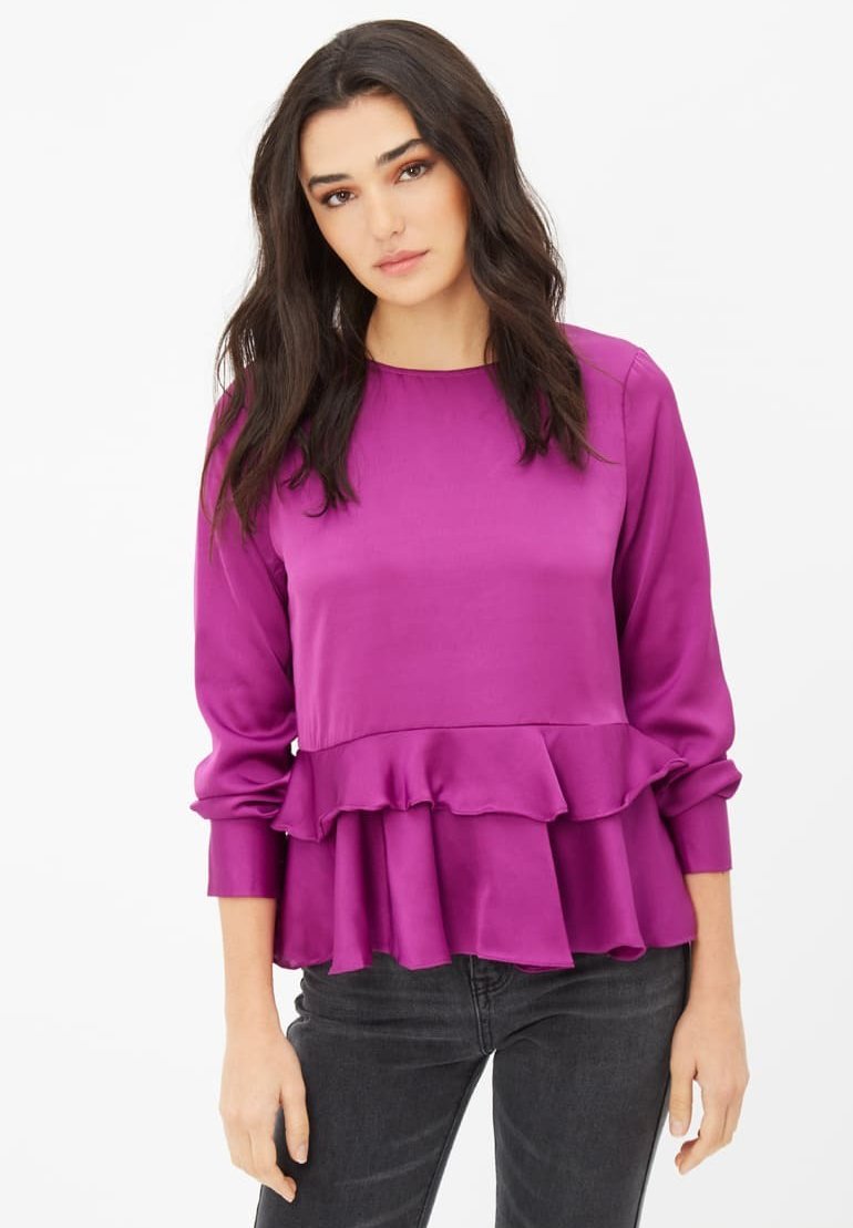 Blusa fucsia de tela suave con mangas largas, que presenta un bajo con volantes y un escote recto. Combinada con unos vaqueros oscuros.