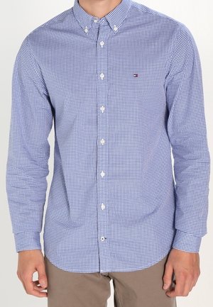 Mann trägt ein blau-weiß kariertes, langärmeliges Hemd mit Knöpfen und beige Hosen, stehend vor einem schlichten Hintergrund.