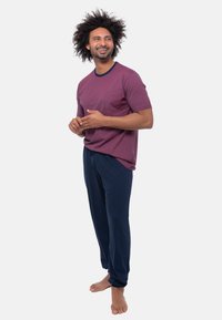 Glunderende man met krullend haar staat blootsvoets in een bordeauxgestreept shirt en marineblauwe broek, en houdt zijn handen samen in een ontspannen houding.