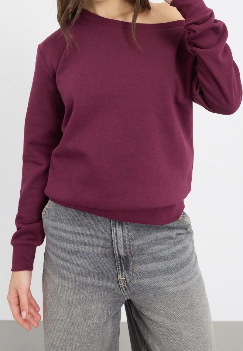 Sweat-shirt bordeaux à épaules dénudées en tissu doux avec manches longues, poignets côtelés et coupe décontractée, associé à un jean gris délavé.