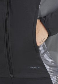 Veste de sport noire avec fermeture éclair, au design épuré et accents gris à motifs, fabriquée dans un matériau lisse et extensible.