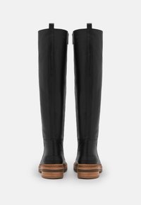 Schwarze kniehohe Lederstiefel mit braunen gestapelten Absätzen, sichtbaren Rückennähten und Zuglaschen oben, von hinten auf einem weißen Hintergrund präsentiert.