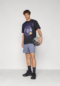 Sort t-shirt med lilla "Lakers" grafik, parret med lyseblå shorts. Emnet holder en grå basketball, iført sorte sko og sokker.