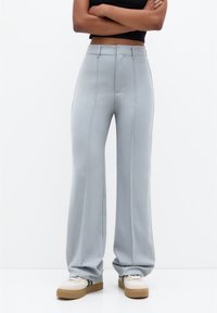 Personne portant un pantalon large taille haute gris clair et des baskets blanches avec semelles beige, debout devant un fond blanc uni.