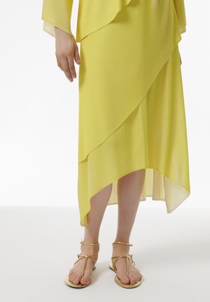 Robe jaune plissée avec un ourlet asymétrique, design superposé et sandales dorées à lanières sur pieds nus, sur fond blanc.