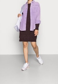 Lila kordflöjelsskjorta över en mörkbrun klänning, kombinerad med vita och rosa sneakers. En ljusgrå väska hänger över axeln.