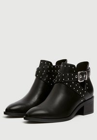 Botas de cuero negras hasta el tobillo con puntera puntiaguda, detalle de correa con tachuelas, hebilla ajustable y un tacón bajo de bloque. Textura suave con acentos.