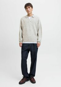Ung mand iført en lys beige strikket sweater, mørkeblå jeans, brune lædersko, stående foran en ensfarvet hvid baggrund.