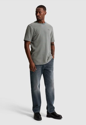 Homme debout portant une chemise à manches courtes rayée gris et blanc, un jean bleu, des chaussures noires, une main dans la poche, regardant vers sa gauche.